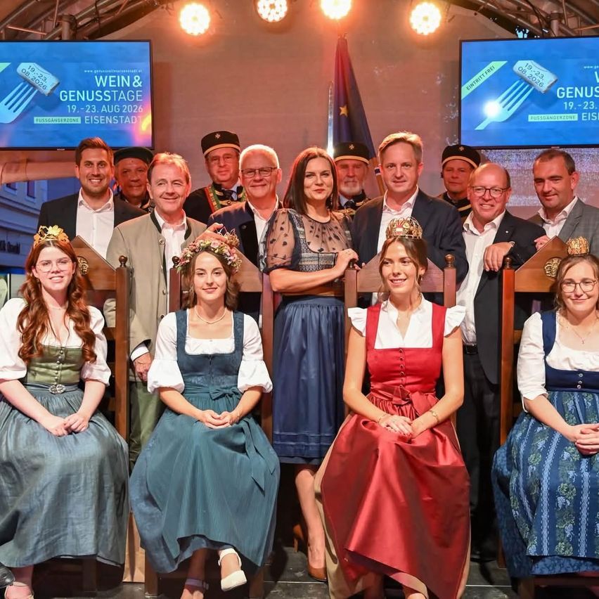 Eine Gruppe von Menschen in traditioneller deutscher Kleidung, einschließlich Dirndl und Lederhosen, posiert für ein Foto auf einer Bühne mit einem Werbebanner für das Wein & Genuss Festival. Die Veranstaltung findet in Eisenstadt statt.