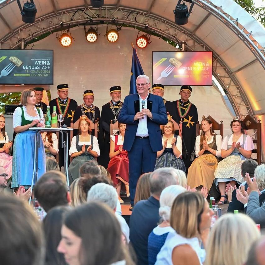 Eine Gruppe von Menschen in traditioneller bayerischer Kleidung steht auf der Bühne, klatscht und lächelt. Ein Mann in einem blauen Anzug spricht in ein Mikrofon. Hinter ihm befinden sich zwei große Bildschirme mit dem Text 'Wein & Genussstage'.