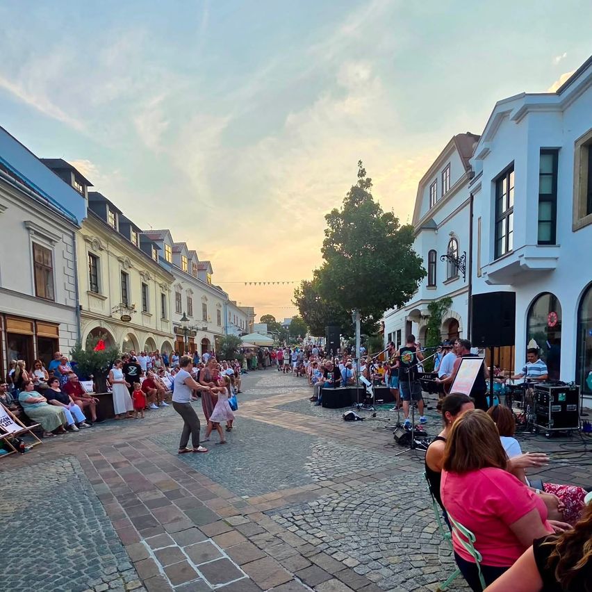 Eine Straßenaufführung mit einer Band in der Mitte einer Kopfsteinpflasterstraße, umgeben von einer Menge Menschen, die auf Stühlen sitzen und stehen.