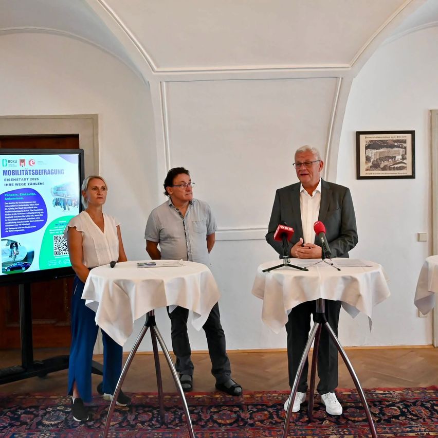 Drei Personen stehen an einem Podium mit Mikrofonen, ein großer Bildschirm mit 'Mobilitätsbefragung' hinter ihnen. Der Mann rechts trägt einen Anzug, der Mann in der Mitte ein helles Hemd und die Frau links eine weiße Bluse.