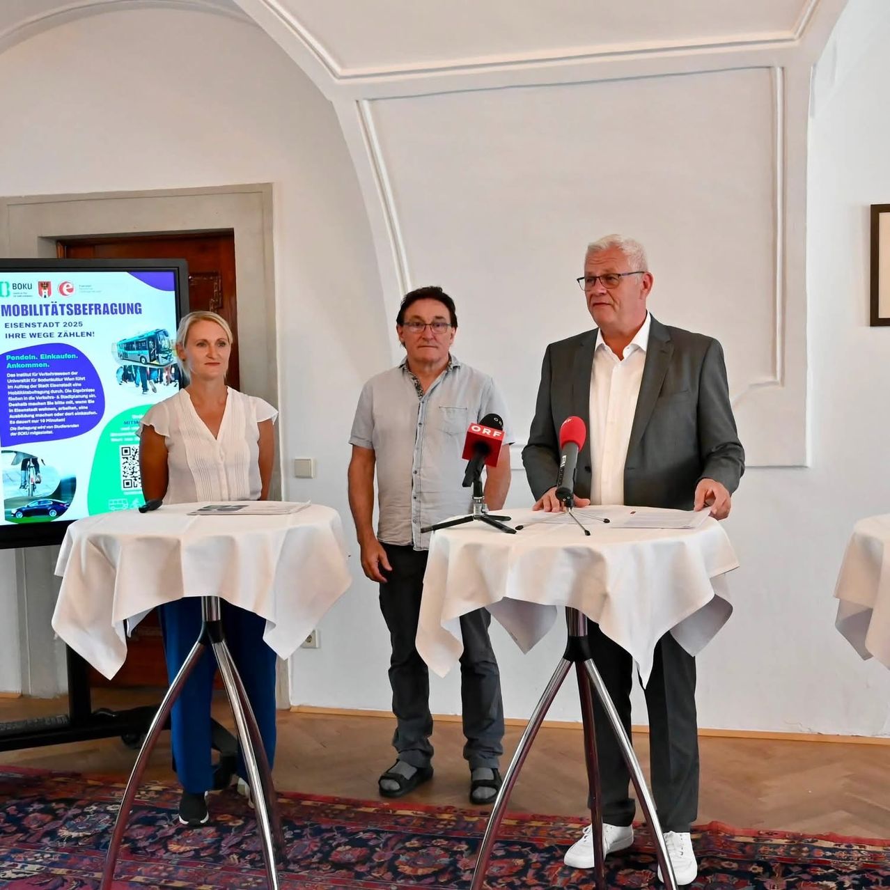 Drei Personen stehen an einem Podium mit Mikrofonen vor einer Leinwand, die einen Bus zeigt. Eine Frau in einem weißen Shirt und blauer Hose steht links, ein Mann in einem hellblauen Shirt und schwarzer Hose in der Mitte und ein Mann in einem grauen Anzug rechts. Dahinter befindet sich eine weiße Wand mit einem großen Bogen.