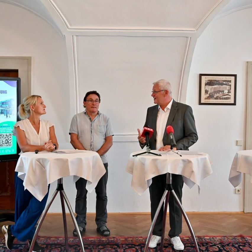 Drei Personen stehen hinter einem Tisch, zwei Frauen und ein Mann, mit Mikrofonen vor ihnen. Der Mann spricht, während die anderen zuhören. Hinter ihnen ist eine Wand mit einem gerahmten Bild und einem Monitor.