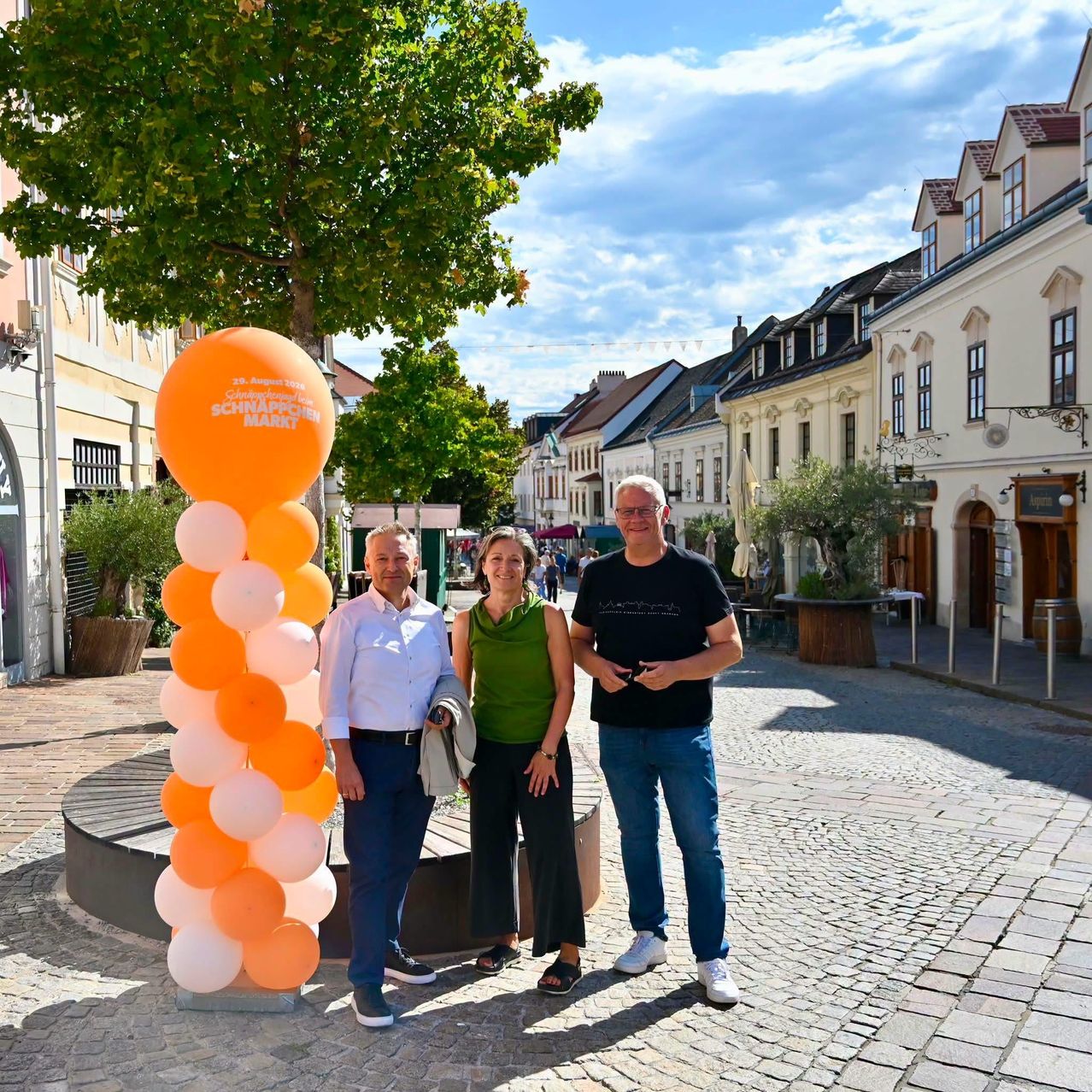 Drei Personen stehen vor einer großen Ballondekoration auf einer Kopfsteinpflasterstraße mit Gebäuden und Bäumen im Hintergrund.