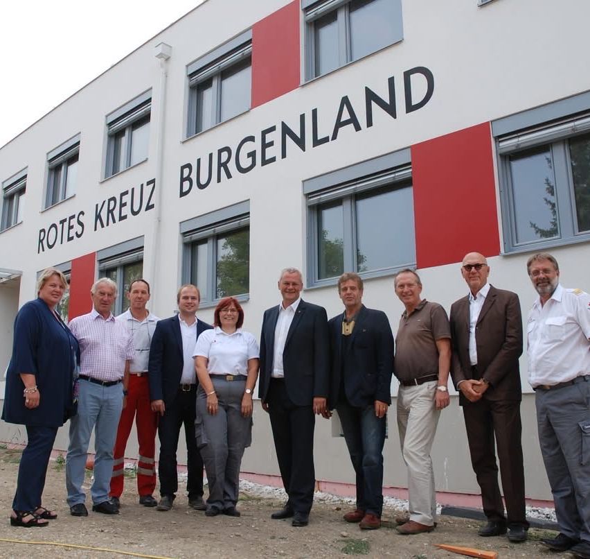 A group of people in formal attire stand in front of a building with the text 'Rotes Kreuz Burgenland' on the wall.