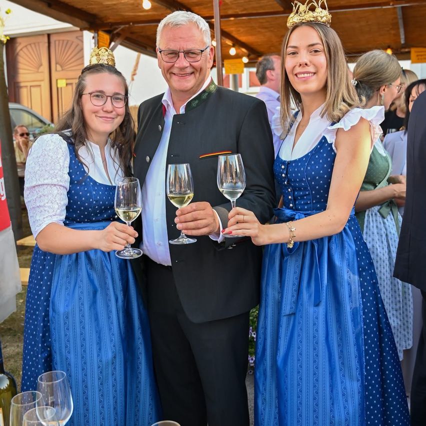 Drei Personen in traditioneller Kleidung, darunter ein Mann in Anzug, halten Weinglas. Sie stehen vor einer Holzstruktur.