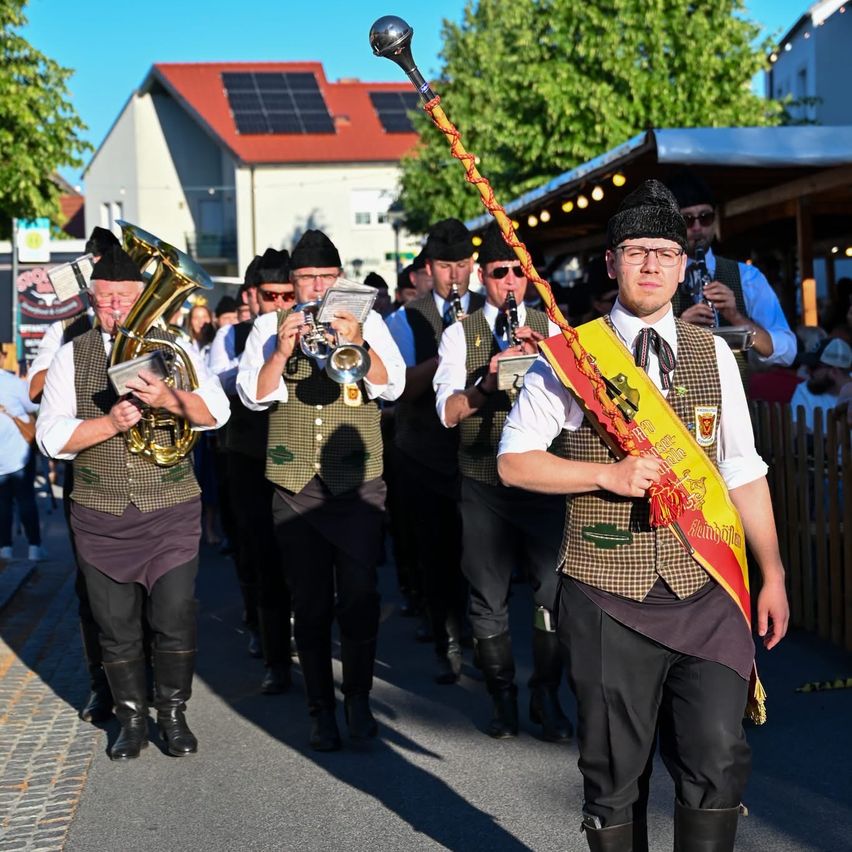 Eine Blaskapelle zieht in traditioneller Kleidung die Straße entlang. Einige spielen Blechblasinstrumente. Ein Gebäude mit Solarzellen befindet sich dahinter.