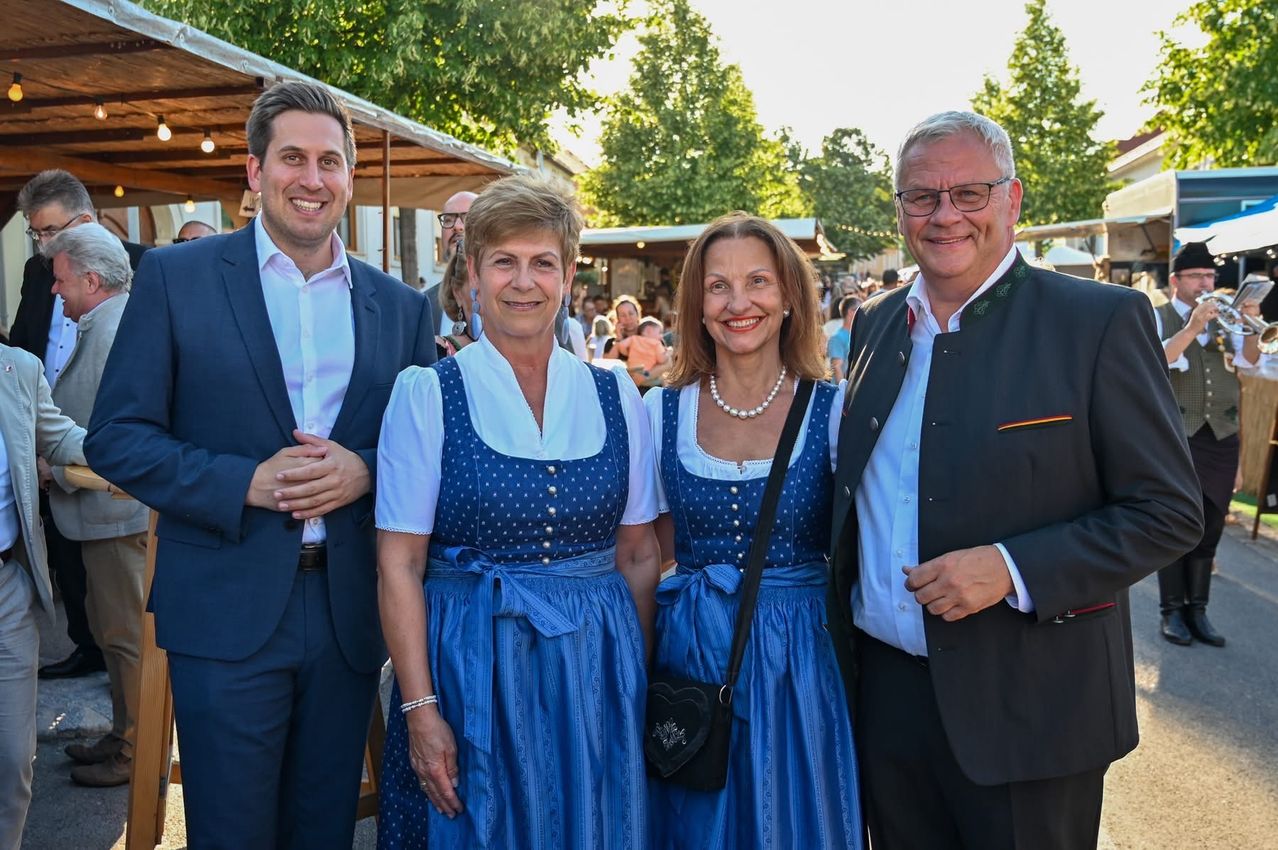Vier Menschen posieren für ein Foto bei einer Outdoor-Veranstaltung. Zwei Frauen tragen passende blaue Kleider und die Männer formelle Kleidung. Bäume und eine Menschenmenge sind im Hintergrund sichtbar.
