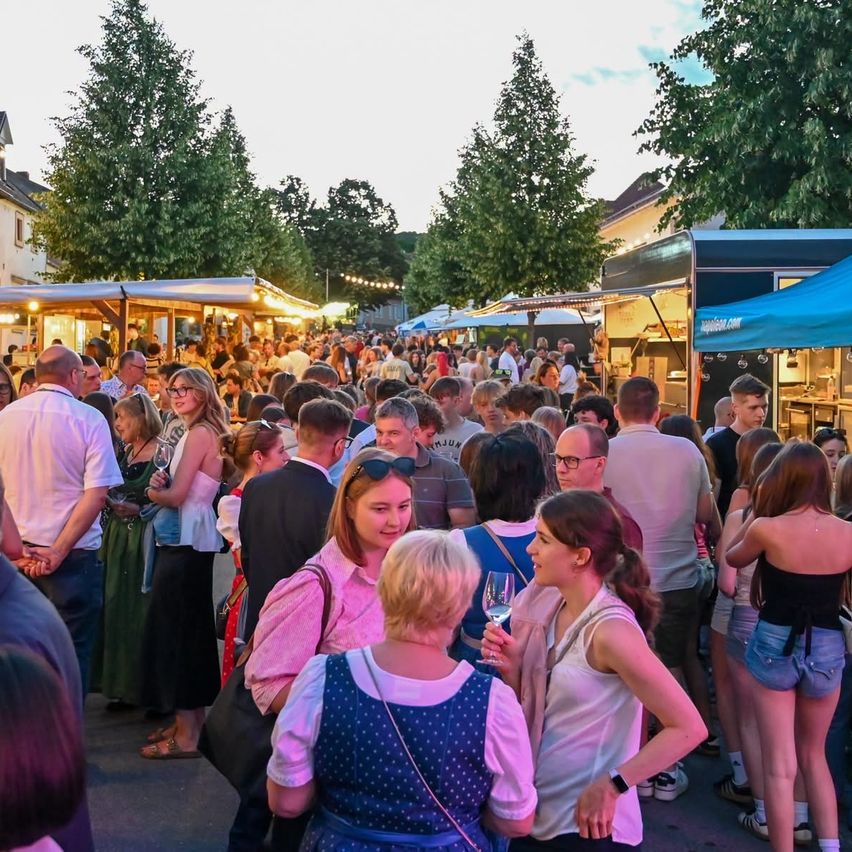 Menschenmengen stehen in einer Schlange an Imbisswagen, unter einem blauen Zelt, auf einem Stadtplatz. Bäume und Gebäude sind im Hintergrund zu sehen.