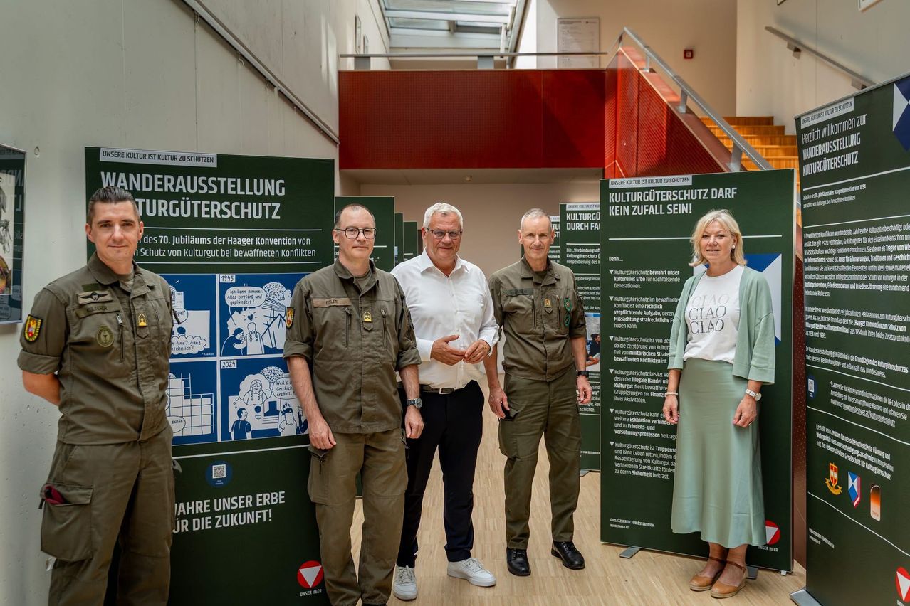 Fünf Personen stehen vor einer grünen Ausstellung, drei Männer in Militäruniformen und zwei Männer in Zivilkleidung. Sie scheinen für ein Foto zu posieren. Die Ausstellung hinter ihnen hat Text und Bilder.