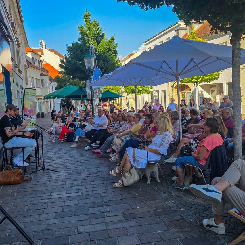 Ein Mann spielt Gitarre auf einer Kopfsteinpflasterstraße, während ein Publikum auf Stühlen und unter Schirmen sitzt. Gebäude und Bäume säumen die Straße.
