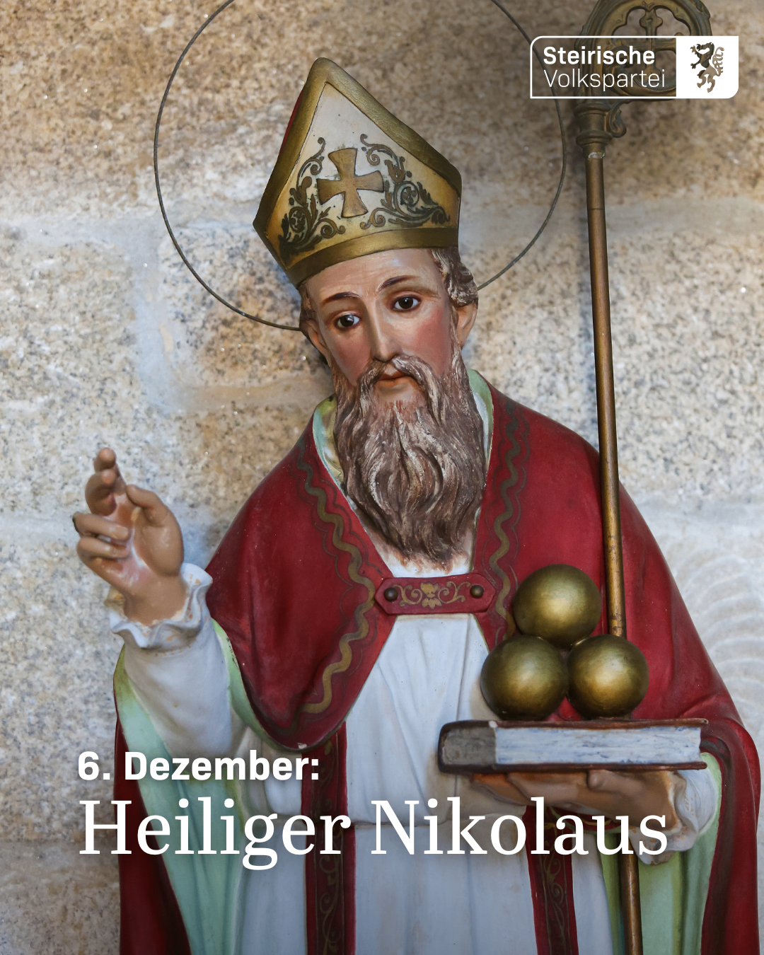 Eine Statue des Heiligen Nikolaus, gekleidet in einen roten Umhang und mit einem Buch und drei goldenen Kugeln in der Hand, vor einer Steinmauer. Der 6. Dezember ist in deutscher Sprache über der Statue geschrieben.