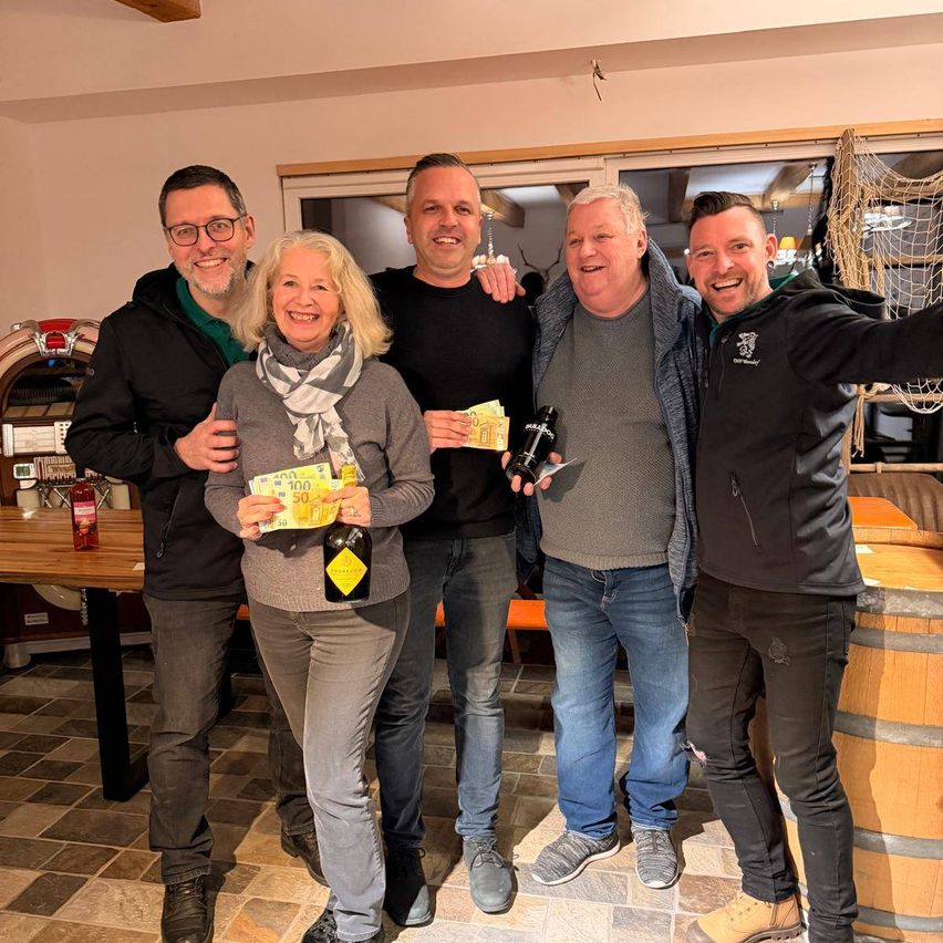 Fünf Erwachsene posieren für ein Foto in einem Raum mit Fass, Tisch und Jukebox. Eine Frau hält eine Flasche und Bargeld, und ein Mann hält eine Flasche.