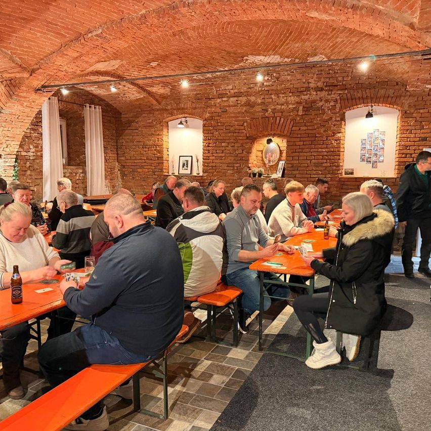 Eine Gruppe von Menschen sitzt in einem Restaurant mit Ziegelwänden um Tische herum und spielt Karten. Die Tische sind mit orangefarbenen Tüchern bedeckt, und Flaschen Bier stehen darauf.