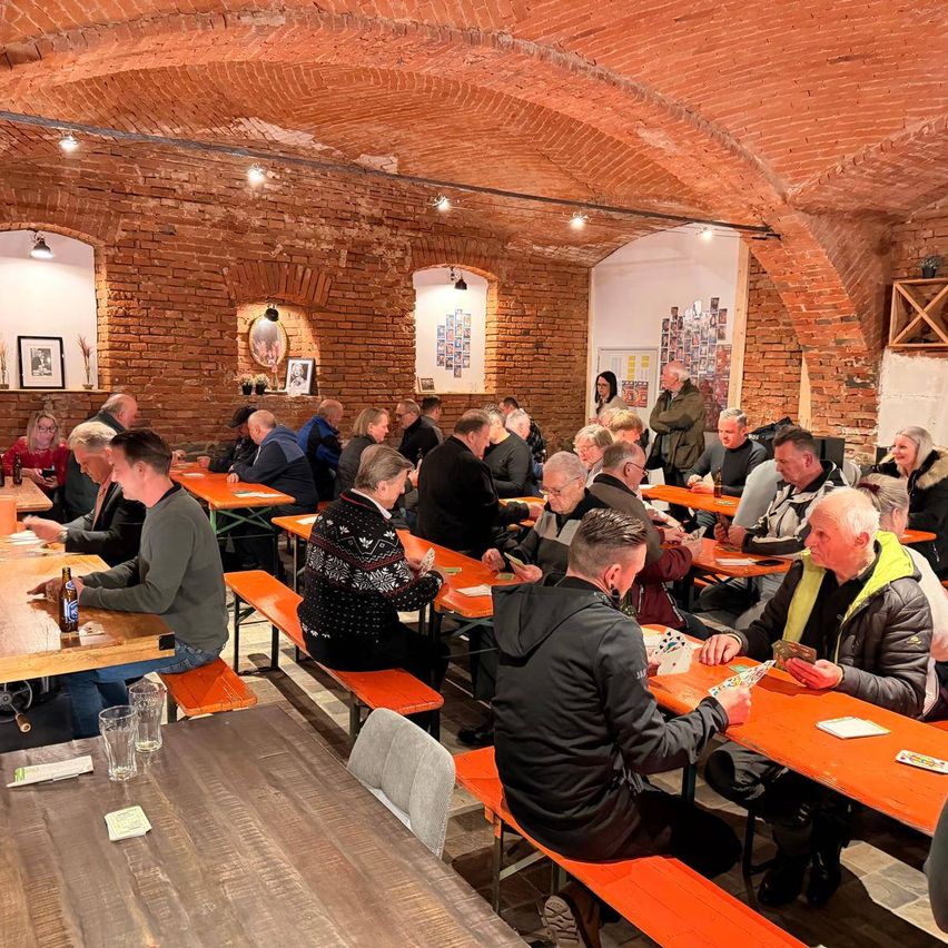 Eine Gruppe von Menschen sitzt an orangefarbenen Bänken um Holztische in einem kellerartigen Raum mit Ziegelwänden. Einige spielen Karten, während andere sich unterhalten. Flaschen und Gläser stehen auf den Tischen. Die Wände sind mit gerahmten Bildern und einer Topfpflanze geschmückt.