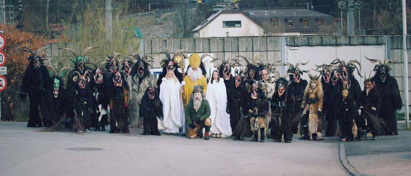 Eine Gruppe von Menschen in Kostümen steht vor einem Gebäude mit einem Zaun und einem abschüssigen Hügel. Die Kostüme umfassen eine Weihnachtsmannfigur, einen Mann in einem grünen Mantel und mehrere andere in schwarzen Kostümen mit Masken.