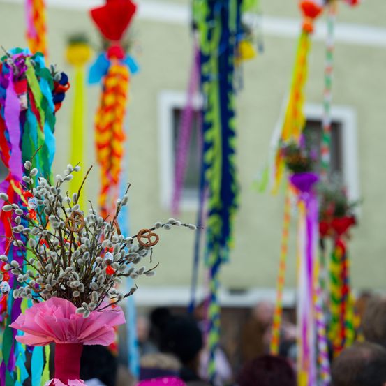 Ein Nahaufnahme einer bunten Weidenzweige, dekoriert mit bunten Bändern und Brezeln, steht im Zentrum einer Menschenmenge. Der Hintergrund ist unscharf, mit Menschen in der Ferne und einem grünen Gebäude.