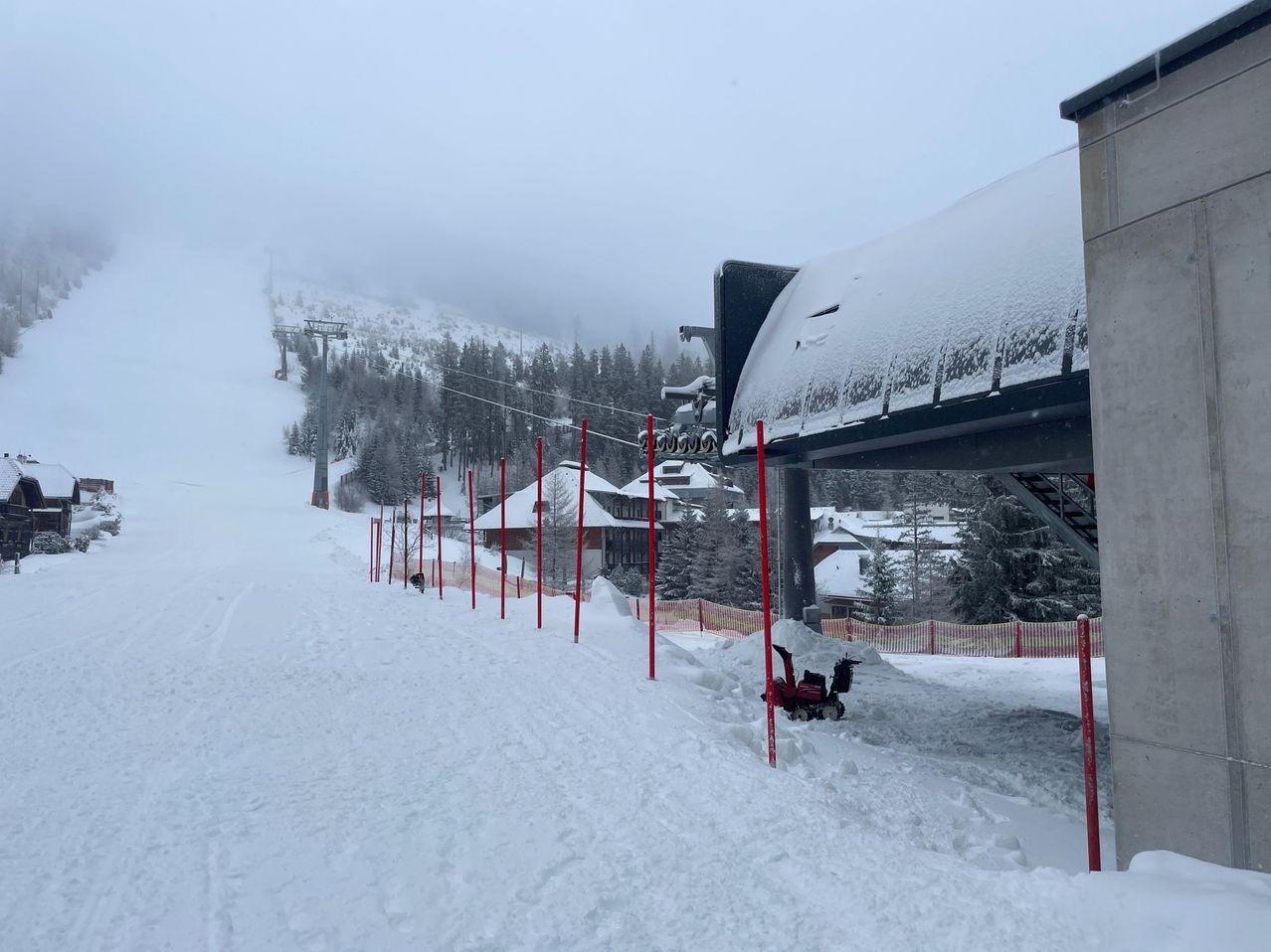 Eine verschneite Skipiste mit einem roten Schneepflug und roten Stangen, Häusern und Bäumen im Hintergrund, die ebenfalls mit Schnee bedeckt sind.