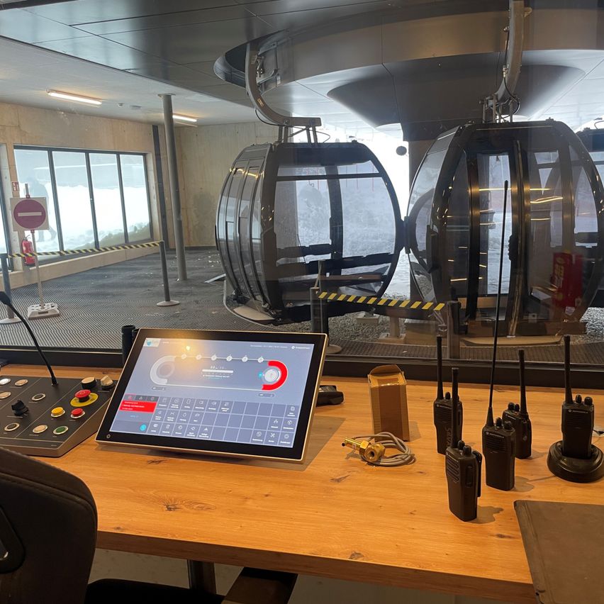 Ein Kontrollraum mit einem Monitor, der eine Bedienoberfläche anzeigt, mehrere Walkie-Talkies und einem Tisch mit Tastatur. Zwei Seilbahnkabinen sind vor dem Glasfenster geparkt.