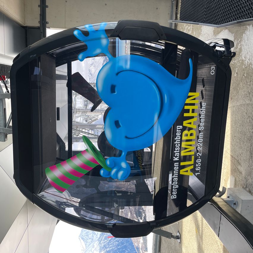 Das Bild zeigt eine Gondel mit einer blauen Comicfigur und Text auf Deutsch. Die Comicfigur hält einen bunten gestreiften Kegel. Die Gondel hat Glasfenster.