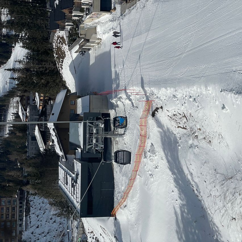 Eine Luftaufnahme eines Skigebiets mit einer Gondelbahn und Skifahrern auf der Piste. Mehrere Personen stehen auf dem schneebedeckten Boden, einige in roten Jacken. Das Gebiet ist von Bäumen und Gebäuden umgeben.