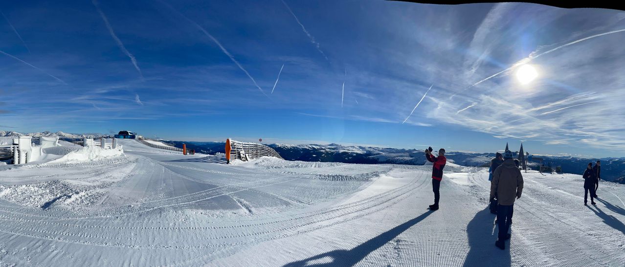 Eine Person steht auf einem schneebedeckten Berggipfel und fotografiert den klaren blauen Himmel mit Kondensstreifen. Der Schnee ist frisch präpariert, und in der Ferne erheben sich Berge.
