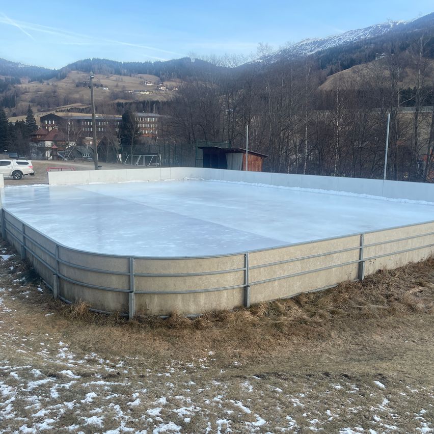 Eine leere Eisbahn mit einer Betonbarriere ist von einem kahlen Feld mit Schnee umgeben. In der Ferne befinden sich Häuser, ein Gebäude und eine Bergkette.