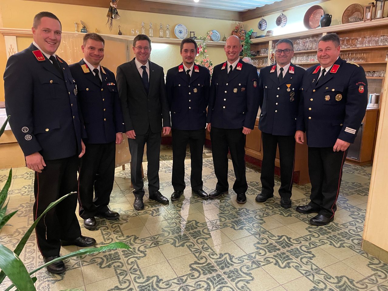 Eine Gruppe von Männern in Uniformen steht in einem Raum. Sie lächeln und posieren für ein Foto. Der Boden ist gefliest.