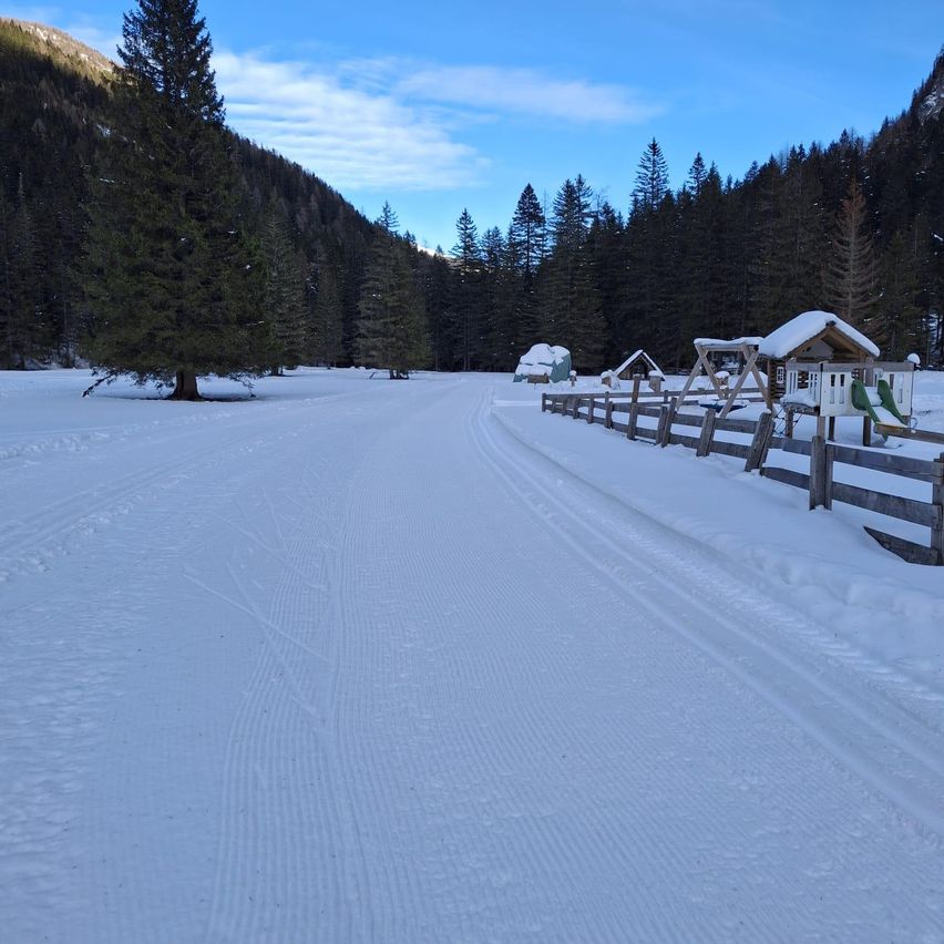 Eine verschneite Landschaft mit einem Holzzaun und einem Spielhaus mit einer Rutsche. Der Weg ist klar und umgeben von Kiefern und Bergen unter einem blauen Himmel.