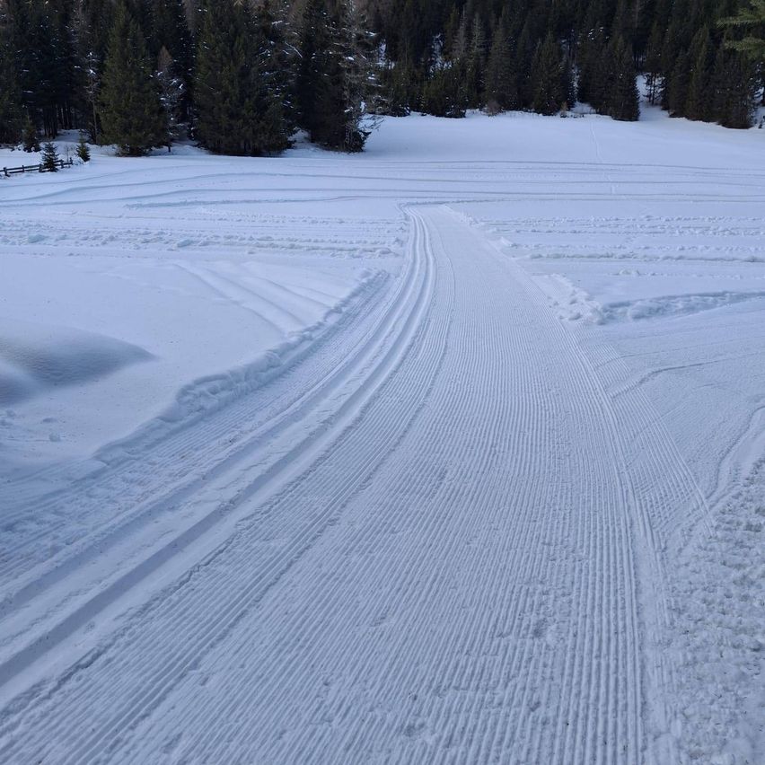 Eine verschneite Landschaft mit einem gut definierten Pfad. Der Pfad ist mit Spuren bedeckt, wahrscheinlich von Fahrzeugen oder Skiern. Der Schnee ist gleichmäßig auf dem Boden verteilt. Bäume säumen den Horizont und schaffen eine friedliche Winterszene.