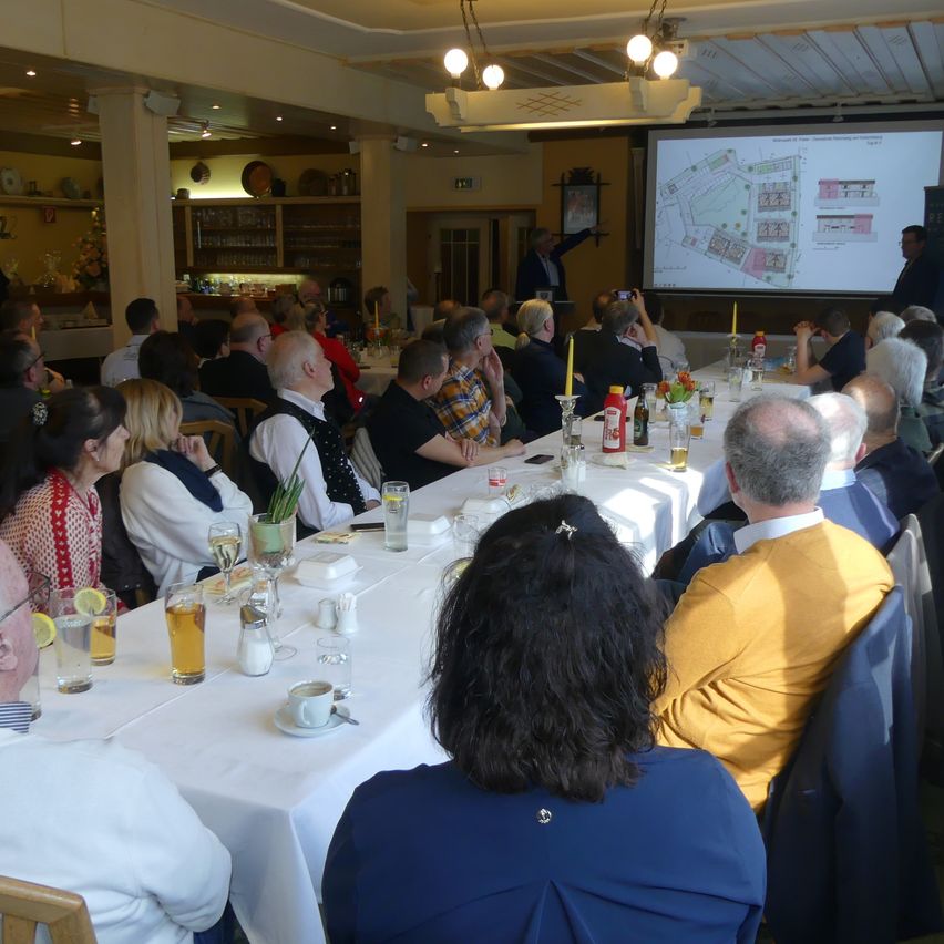 In einem großen Raum wird eine Konferenz abgehalten, wobei eine Projektionsleinwand einen Grundriss zeigt. Die Teilnehmer sitzen an langen Tischen mit Getränken und Snacks. Ein Mann präsentiert vorne.