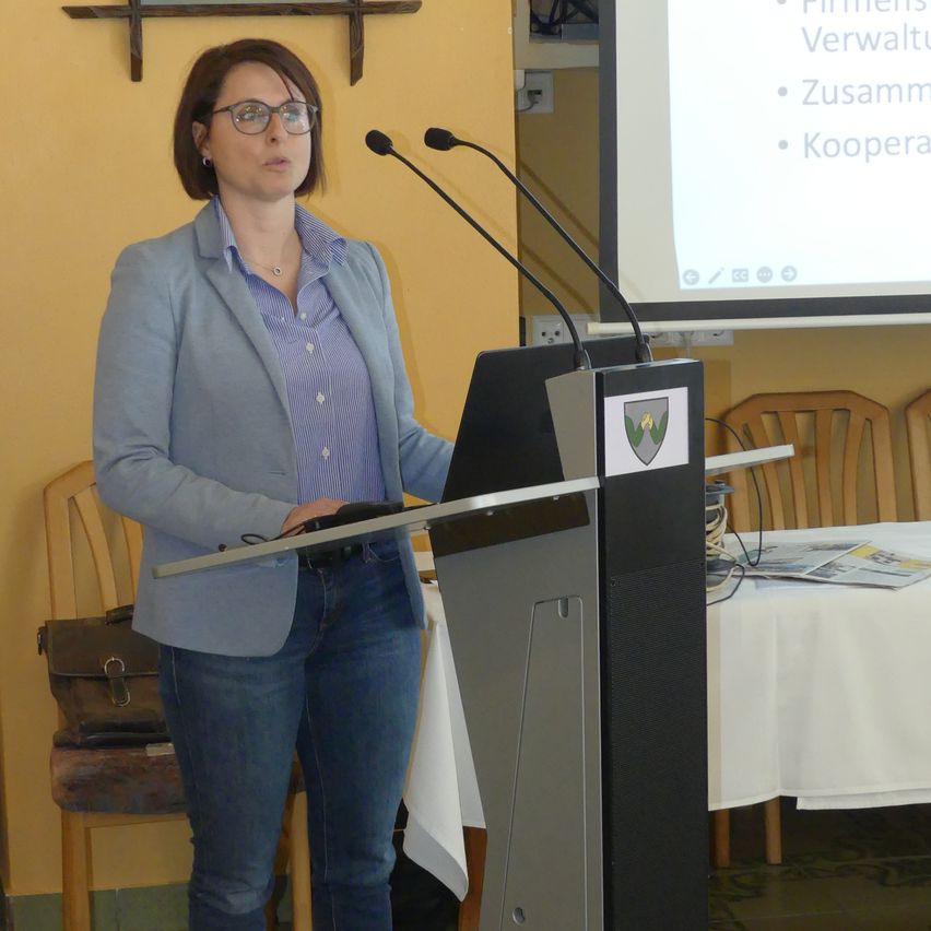 Eine Frau steht an einem Podium mit Mikrofonen vor einer Projektionsleinwand mit Aufzählungspunkten, trägt einen blauen Blazer und eine Brille.