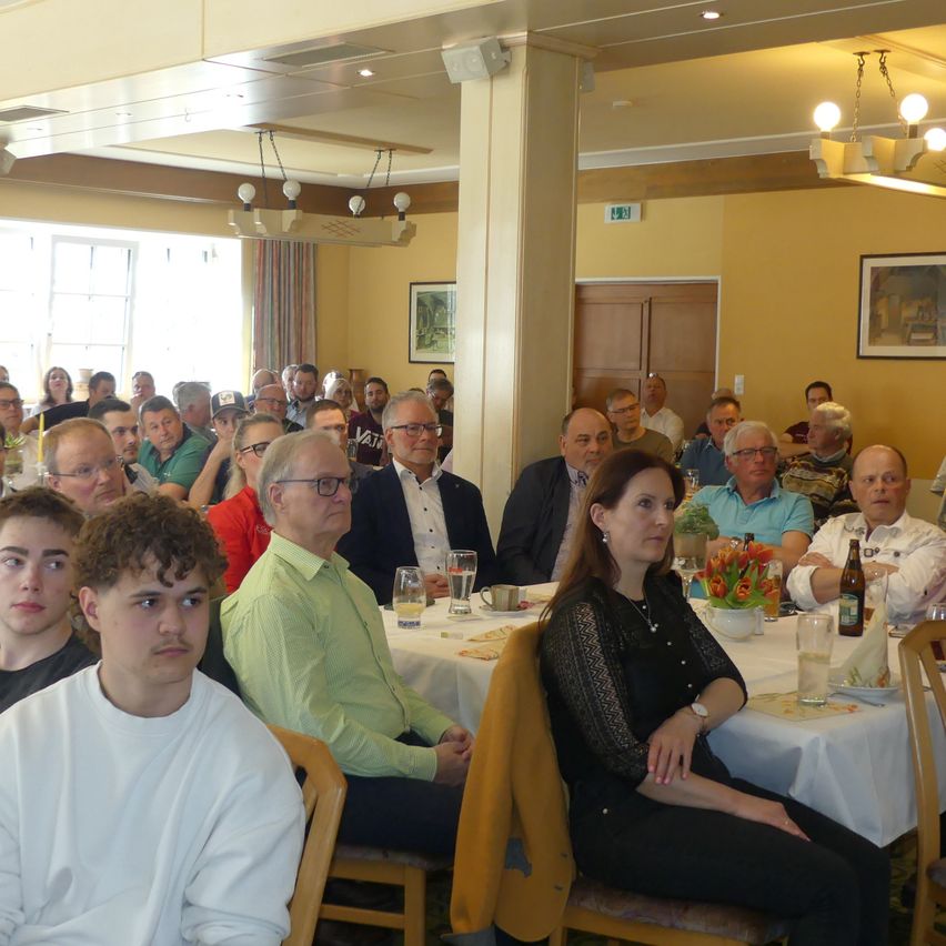Eine Gruppe von Menschen, darunter Männer und Frauen, sitzt an einem Tisch. Der Tisch ist mit einem weißen Tuch bedeckt und hat eine Flasche, einen Becher und einen Teller darauf. Die Menschen schauen in die gleiche Richtung. In der Mitte des Raumes befindet sich eine Säule.