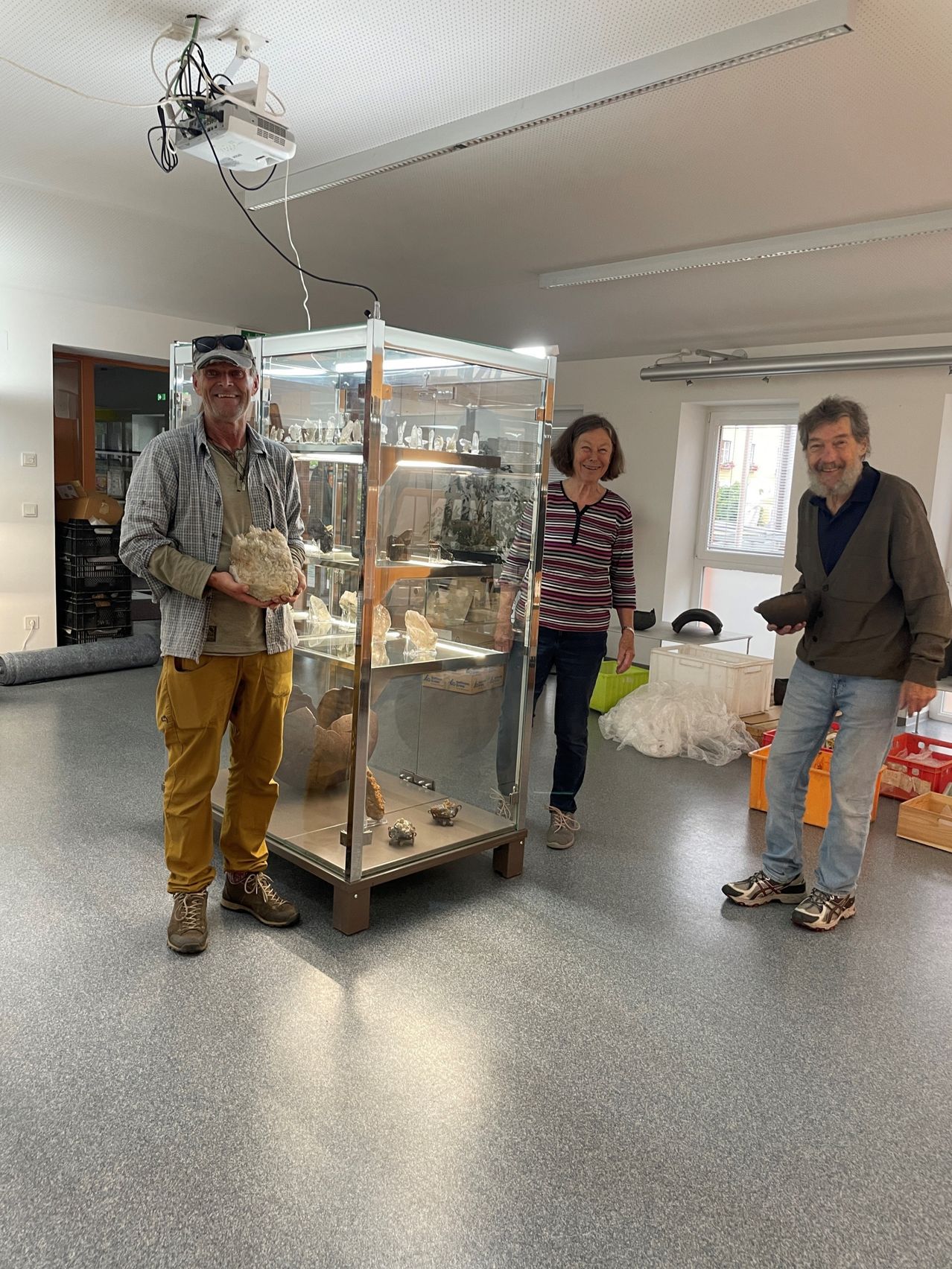 Drei Menschen stehen neben einer Glasvitrine in einem Raum. Ein Mann hält einen großen Felsen. Ein anderer hält ein Artefakt. Eine Frau schaut zur Kamera.