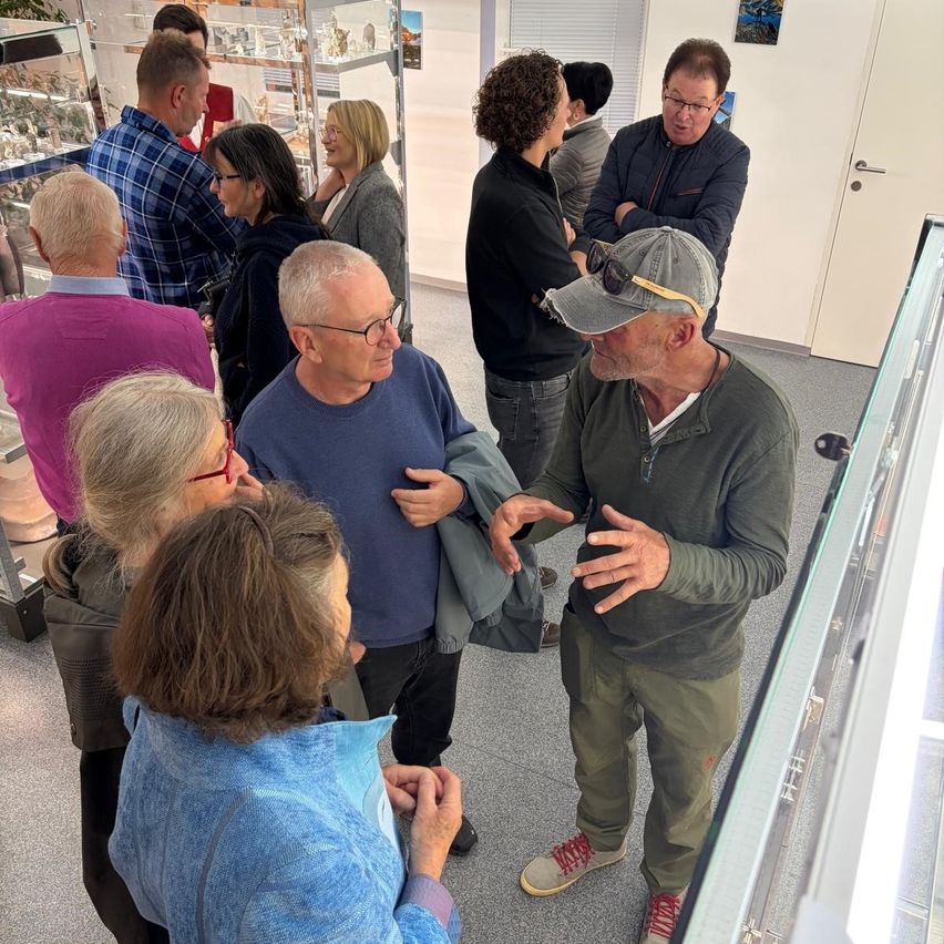 Eine Gruppe Erwachsener ist in einem Museum und diskutiert. Ein Mann mit Hut und Sonnenbrille spricht mit einer Frau. Hinter ihnen schauen andere Leute Gegenstände in der Ausstellung an.