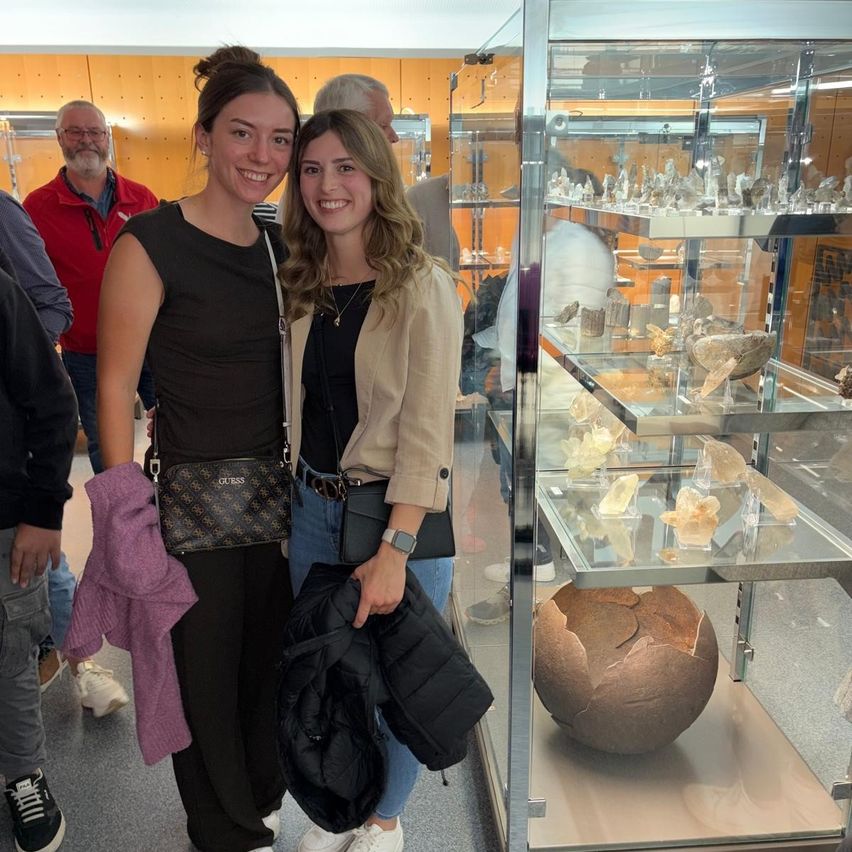 Zwei Frauen stehen lächelnd vor einer Glasvitrine mit Mineralien und einem Fossil. Hinter ihnen betrachten einige Leute ebenfalls die Gegenstände in der Vitrine.