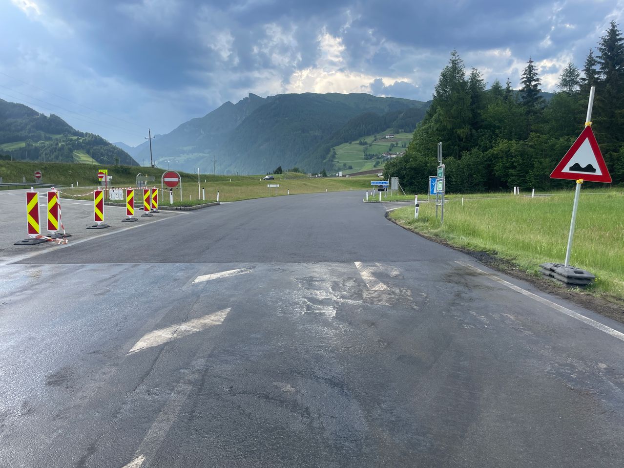Eine leere Straße mit Verkehrszeichen und Absperrungen, umgeben von grünem Gras und Bergen im Hintergrund.