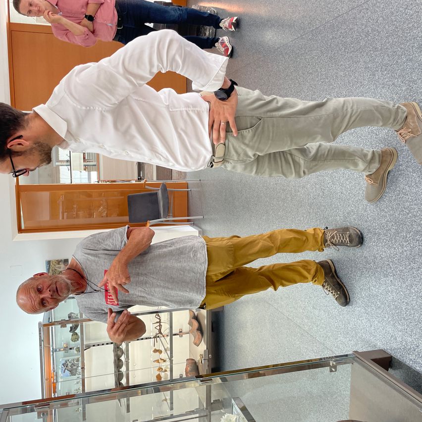 Ein Mann in einem weißen Hemd und khakifarbener Hose beugt sich vor, während ein anderer in einem grauen Hemd und gelben Hosen steht und ein rotes Schild trägt. Dahinter steht eine Person in einem rosa Hemd. Dahinter ist eine Vitrine mit Objekten darin.