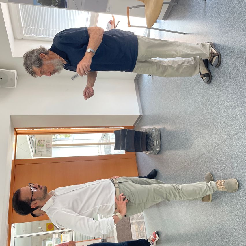 Zwei Männer stehen in einem Innenbereich, möglicherweise in einem Flur. Der Mann auf der linken Seite zeigt auf etwas, während der andere Mann ihn ansieht. Beide sind lässig gekleidet. Ein Stuhl und ein Lautsprecher befinden sich hinter ihnen, und eine Glastür ist auf der linken Seite.