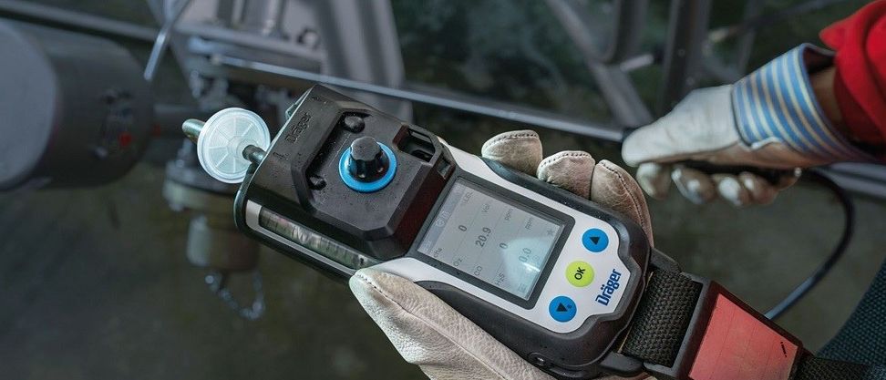 Eine Person mit Handschuhen hält ein Gerät mit Bildschirm, Tasten und einer roten Leuchte, möglicherweise ein Gasdetektor.