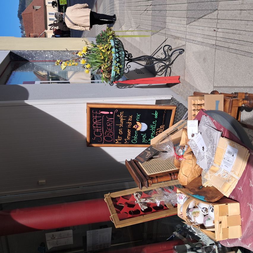 Eine Person geht an einer Blumenarrangement vor einem Geschäft vorbei. Verschiedene Gegenstände sind auf dem Bürgersteig ausgestellt.