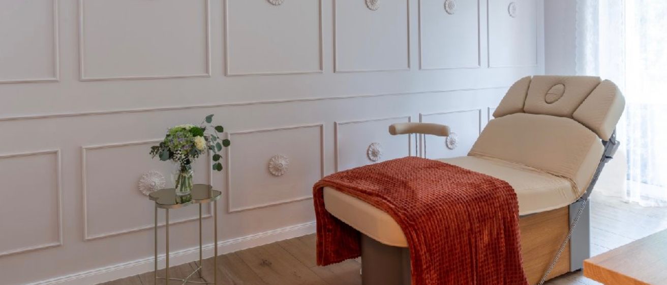 Ein elegantes Zimmer mit einer weißen Wand, die mit Paneelen und einer Kristallleuchte verziert ist. Ein kleiner Tisch hält eine Blumenarrangement, während ein Massagetisch mit einer roten Decke in der Nähe steht.