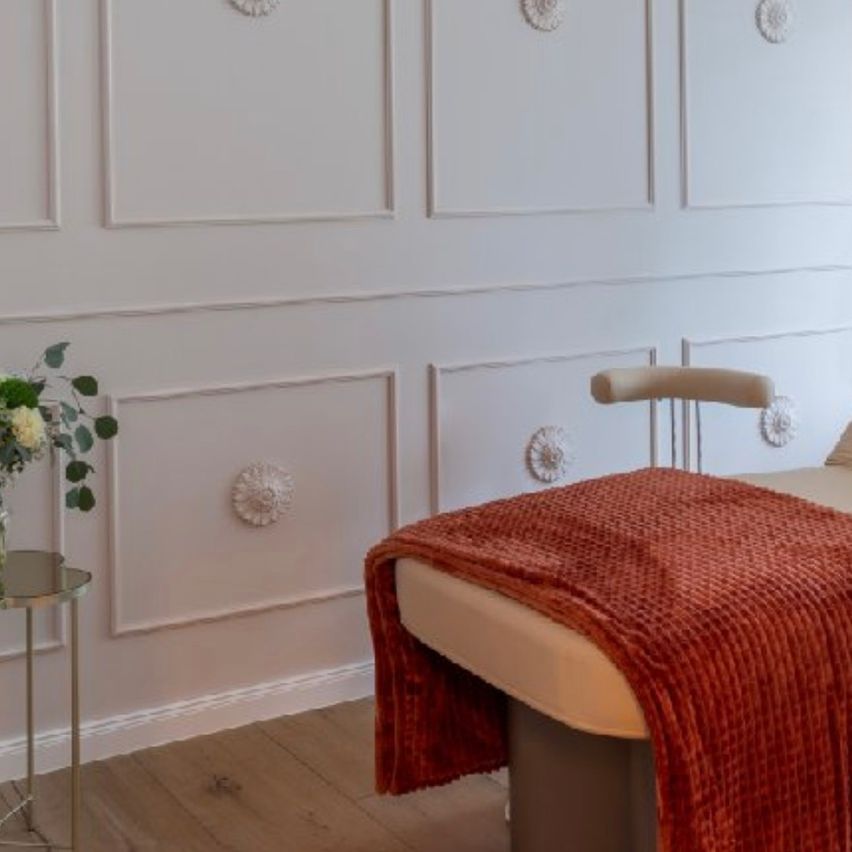 Ein elegantes Zimmer mit einer weißen Wand, die mit Paneelen und einer Kristallleuchte verziert ist. Ein kleiner Tisch hält eine Blumenarrangement, während ein Massagetisch mit einer roten Decke in der Nähe steht.