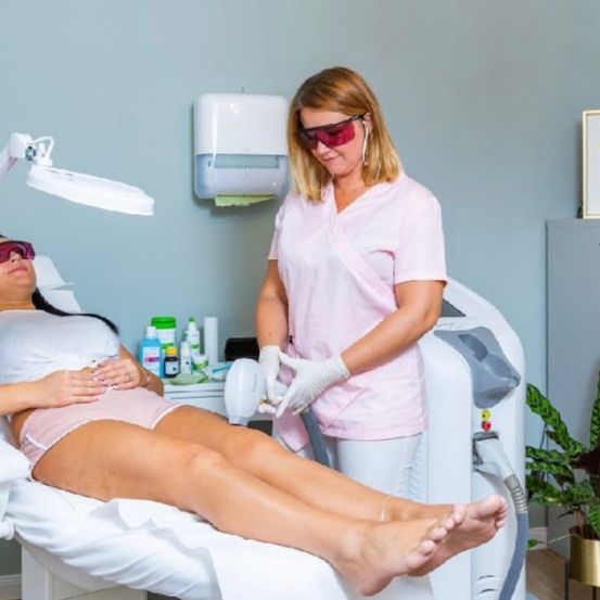 Eine Frau liegt auf einem Stuhl in einem Salon mit einem Gerät, während eine andere Frau steht und ein Gerät in der Hand hält.