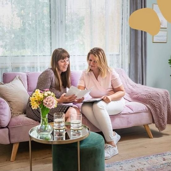 Zwei Frauen sitzen auf einem rosa Sofa und lächeln. Eine liest ein Dokument, die andere schaut zu. Ein Glastisch hält zwei Gläser mit Wasser. Ein Blumenvase steht auf dem Tisch. Hinter ihnen lässt ein Fenster mit weißen Vorhängen Licht herein.
