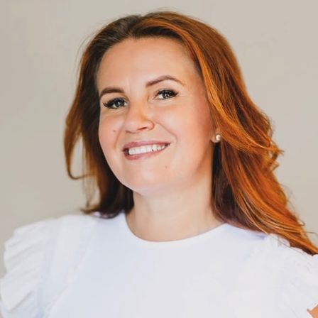 Eine lächelnde Frau mit langen roten Haaren posiert für ein Bild. Sie trägt ein weißes Oberteil mit Rüschen.