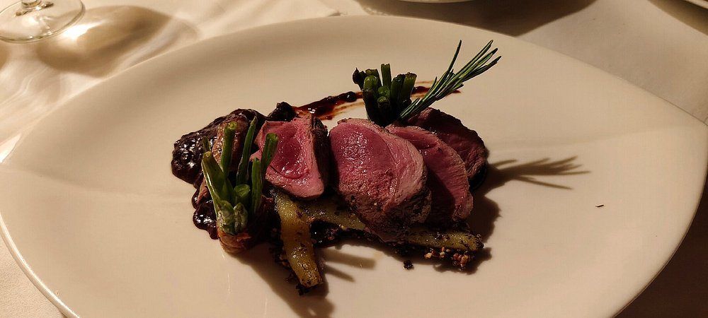 Ein Teller mit roter Sauce, auf dem drei Stücke Fleisch, grüne Bohnen und Rosmarin angeordnet sind, auf einem weißen Teller mit einem leichten Schatten.