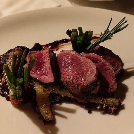 Ein Teller mit roter Sauce, auf dem drei Stücke Fleisch, grüne Bohnen und Rosmarin angeordnet sind, auf einem weißen Teller mit einem leichten Schatten.