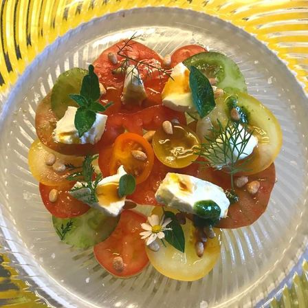 Ein Glasteller mit Tomatensalat, Feta-Käse, Kräutern und Samen auf gelbem Hintergrund.