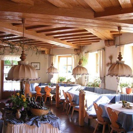 Das Innere eines Restaurants zeichnet sich durch Holzbalken und Möbel aus. Ein Tisch im Vordergrund ist mit Blumen und Flaschen dekoriert. Der Raum hat große Fenster und Vorhänge, und es gibt Pflanzen und Lampen.