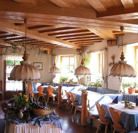 Das Innere eines Restaurants zeichnet sich durch Holzbalken und Möbel aus. Ein Tisch im Vordergrund ist mit Blumen und Flaschen dekoriert. Der Raum hat große Fenster und Vorhänge, und es gibt Pflanzen und Lampen.